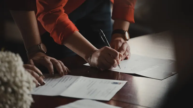 Two people leaning over a dark wooden table, one signing a document with a pen.